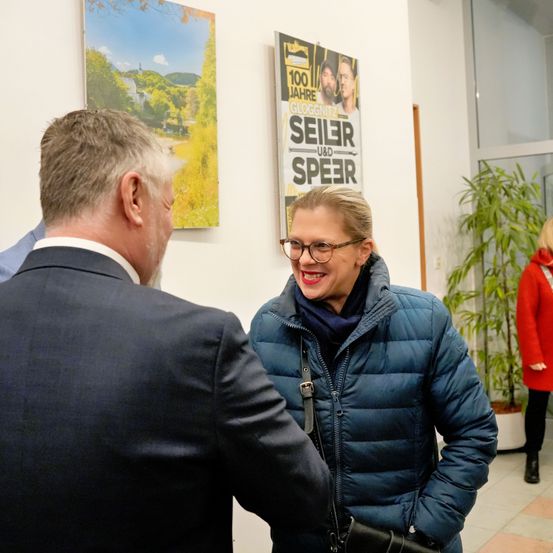 Zwei Personen unterhalten sich in einem Raum mit einem Landschaftsposter und einem Poster mit Logo und dem Text '100 Jahre Gloggnitzer Seiler und Speer'.