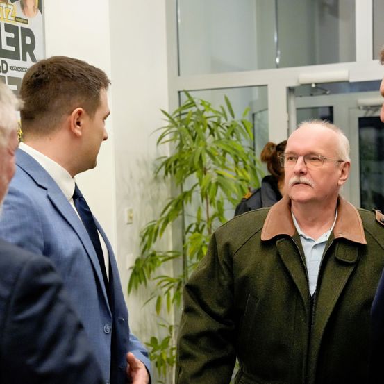 Ein älterer Mann in einem grünen Mantel und mit einer Brille steht vor einer Pflanze und schaut einem Mann in einem blauen Anzug an. Dahinter befindet sich eine Glastür.