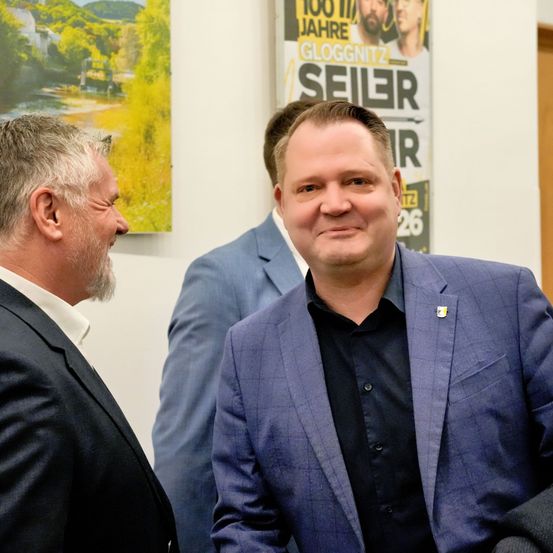 Drei Männer in Anzügen, zwei von ihnen lächelnd, stehen eng beieinander in einem Büro. Hinter ihnen ist ein Plakat mit den Worten Gloggnitzer und Seiler.