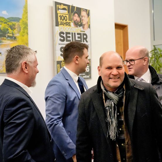 Vier Männer stehen in einem Raum und unterhalten sich wahrscheinlich. Einer trägt eine Mütze und eine Brille. Ein Poster hinter ihnen sagt '100 Jahre Gloggnitzer'.