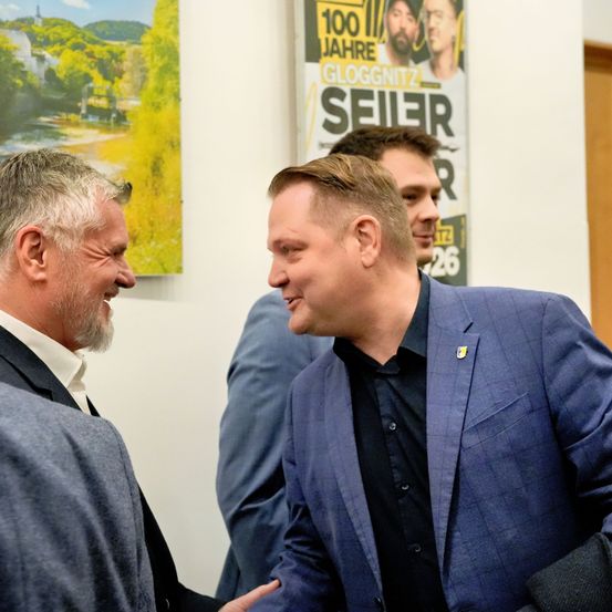 Drei Männer in Anzügen, einer mit einem Abzeichen, stehen in einem Raum, lächeln und unterhalten sich. Hinter ihnen befindet sich ein Plakat mit dem Text 'Gloggnitz 100 Jahre Seiler'.