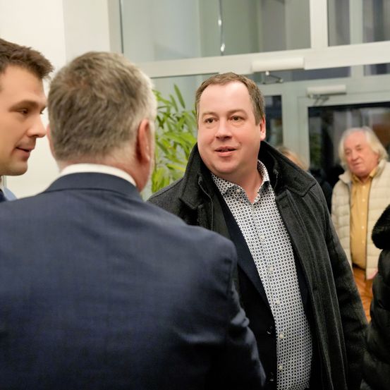 Drei Männer unterhalten sich in einem Raum. Der Mann in der Mitte lächelt und trägt eine schwarze Jacke. Hinter ihm steht ein Mann in einem Anzug. Rechts steht ein Mann mit weißen Haaren. Ein Topf mit einer Pflanze und eine Glastür sind im Hintergrund zu sehen.