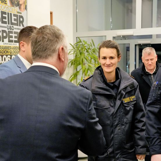 Eine Frau in Uniform lächelt und unterhält sich mit einem Mann im Anzug. Hinter ihnen stehen zwei andere Männer, einer in Anzug und der andere in Mantel. Sie stehen vor einem Plakat und einer Pflanze.