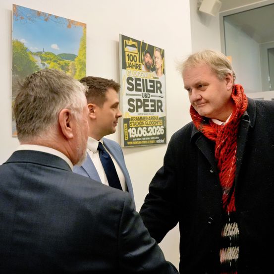 Drei Männer in Anzügen stehen vor einem Plakat. Das Plakat ist für eine Veranstaltung im Stadion Gloggnitz am 19. Juni 2026.