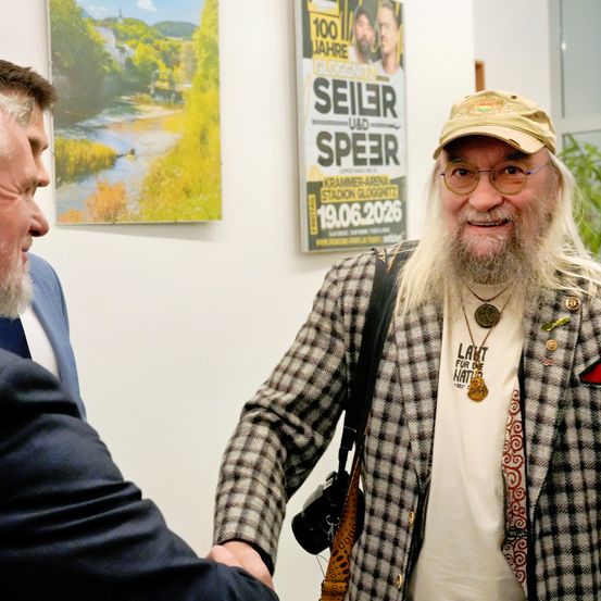 Zwei Männer in einem Raum, einer mit Bart und Brille, trägt eine karierte Jacke und eine Mütze, schüttelt einem anderen Mann im Anzug die Hand. Hinter ihnen ist eine Wand mit einem Plakat, das den Text 'Seilr' und ein Datum zeigt.