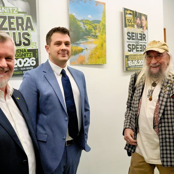 Drei Männer stehen nebeneinander vor einer Wand mit Plakaten und lächeln. Der Mann auf der linken Seite trägt einen schwarzen Anzug, der Mann in der Mitte einen blauen Anzug und der Mann auf der rechten Seite eine karierte Jacke und eine Brille.