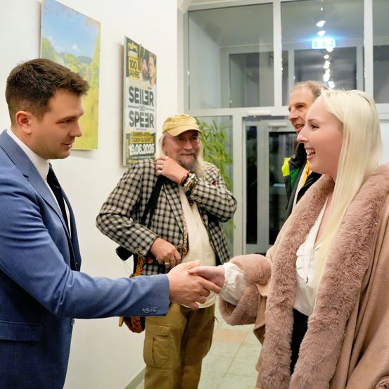 Zwei Personen schütteln sich die Hände in einem Raum. Ein Mann in einem blauen Anzug und Krawatte schaut eine Frau in einem Pelzmantel an. Hinter ihnen stehen zwei Männer, von denen einer eine Tasche hält und einen Hut trägt. Ein Poster mit den Worten 'Seilr und Speer' ist an der Wand angebracht.