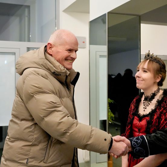 Ein älterer Mann in einer beigen Jacke schüttelt die Hand einer Frau in einem roten und schwarzen Kleid, beide lächeln. Der Ort scheint ein Innenbereich mit Glaswänden und einer weißen Tür zu sein.