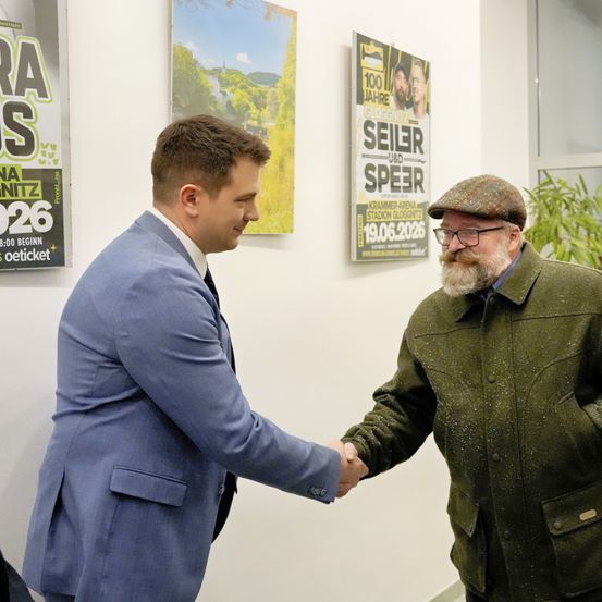 Zwei Männer, einer in einem blauen Anzug und der andere in einer grünen Jacke, schütteln sich die Hände in einem Büro. Hinter ihnen hängen Poster an der Wand, darunter eines für eine Veranstaltung mit Seiler und Speer am 19. Juni 2026.