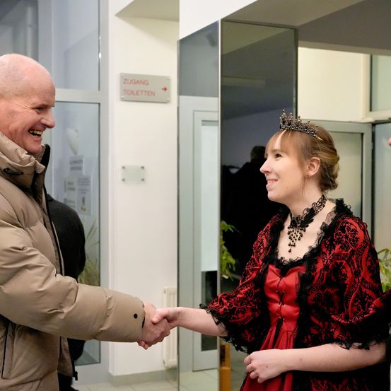 Ein älterer Mann und eine Frau in einem roten Kleid und schwarzem Spitzenschal schütteln sich die Hände. Die Frau trägt eine Krone und eine Halskette. Sie stehen vor einer Glaswand mit einem Schild, das 'Zugang Toiletten' besagt. Hinter ihnen ist ein verschwommenes Bild einer Person.
