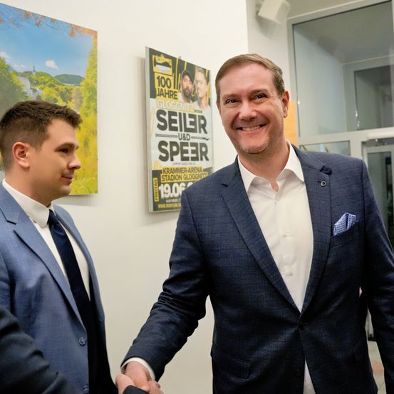 Zwei Männer in Anzügen schütteln sich die Hände in einem Raum mit Postern an der Wand. Ein Poster lautet '100 Jahre Gloggnitzer Seiler und Speir'.