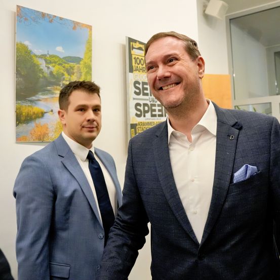 Zwei Männer in Anzügen stehen eng beieinander und lächeln. Der Mann auf der linken Seite trägt einen hellblauen Anzug, während der Mann auf der rechten Seite einen marineblauen Anzug trägt.