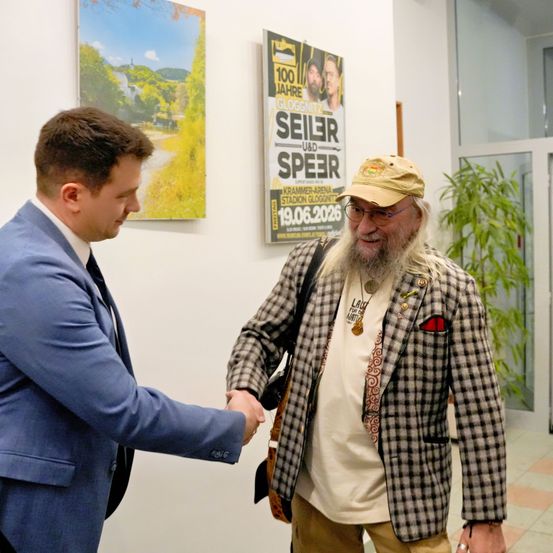 Zwei Männer, einer in einem blauen Anzug und der andere in einem karierten Jackett, schütteln sich die Hände vor einem Poster von Seiler und Speer.