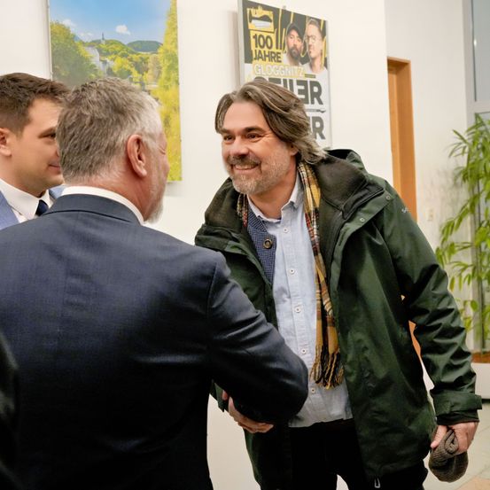 Drei Männer stehen in einem Flur, zwei in Anzügen unterhalten sich mit einem Mann in einer grünen Jacke und einem Schal. Hinter ihnen steht ein Poster mit der Aufschrift 100 Jahre Gloggnitzer.