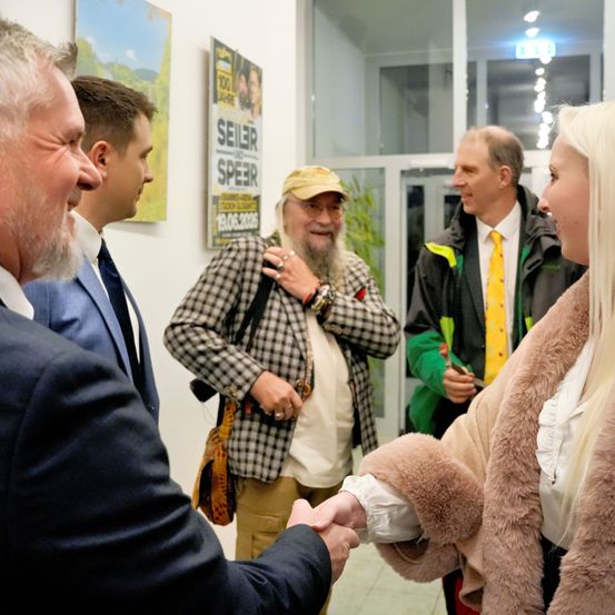 Fünf Personen stehen in einem Raum mit einem Plakat im Hintergrund. Ein Mann schüttelt einer Frau die Hand.