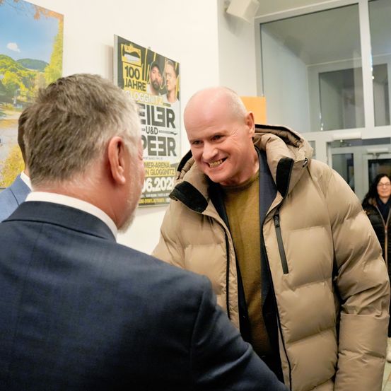 Zwei Männer unterhalten sich in einem Raum, einer in einer braunen Daunenjacke, lächelnd, während der andere in einem blauen Anzug zusieht. Hinter ihnen hängen Poster an der Wand, und eine Frau mit Brille steht in der Nähe der Tür.