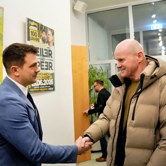 Zwei Männer schütteln sich die Hände in einem Raum mit einem Plakat im Hintergrund. Ein Mann trägt einen Anzug, der andere einen braunen Mantel. Hinter ihnen hält ein Mann ein Telefon.