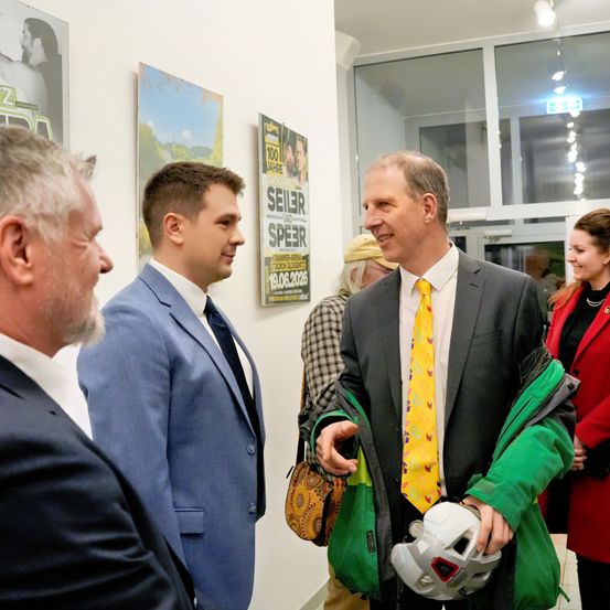 Mehrere Männer und Frauen in Anzügen und Krawatten stehen in einem Innenbereich, wahrscheinlich einer Galerie. Sie scheinen miteinander zu sprechen. Ein Mann hält einen Helm und eine Jacke. Poster hängen an der Wand.