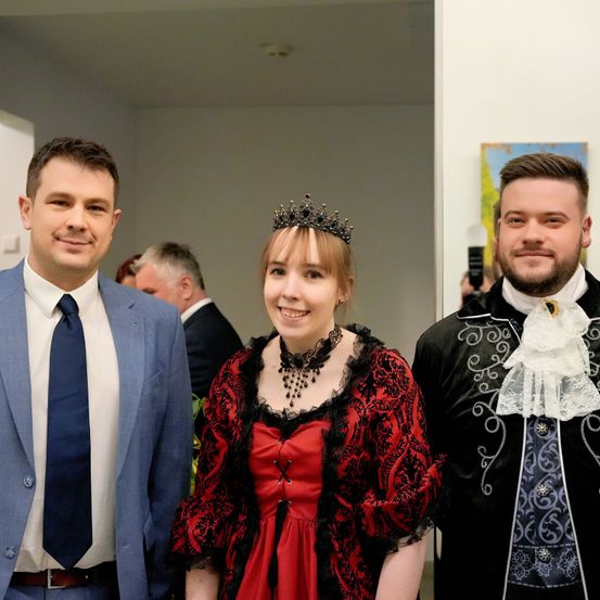 Drei Personen posieren für ein Foto in einem Flur. Der Mann auf der linken Seite trägt einen blauen Anzug und eine Krawatte. Die Frau in der Mitte trägt ein rotes Kleid mit einem schwarzen Spitzenkragen und einer Krone. Der Mann auf der rechten Seite trägt einen schwarzen Mantel mit aufwendigen Designs. Hinter ihnen steht ein weiterer Mann.