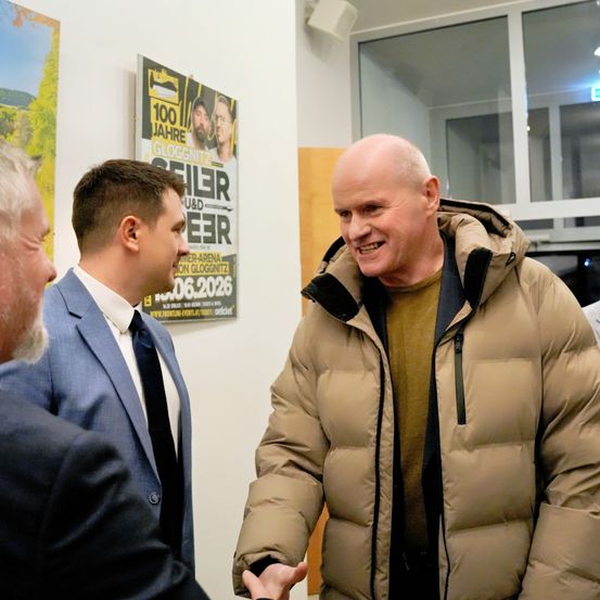 Drei Männer in Anzügen und ein lächelnder Mann in einer beigen Jacke schütteln sich die Hände in einem Raum. Ein Poster mit einem Bild von drei Männern ist an der Wand.