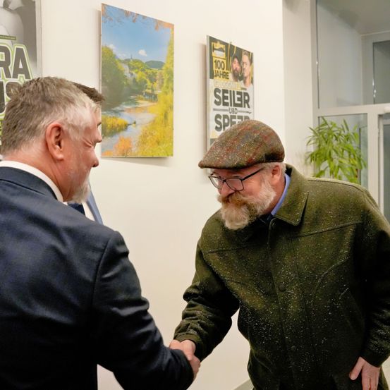 Zwei Männer schütteln sich die Hände in einem Innenbereich. Der Mann auf der rechten Seite trägt einen grünen Mantel und einen Hut. Hinter ihnen befindet sich eine Wand mit Postern.