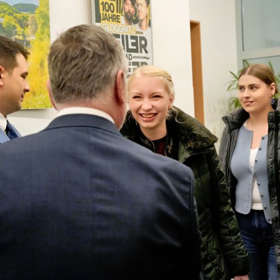 Drei Personen stehen zusammen, zwei Frauen und ein Mann im Anzug, wahrscheinlich in einer Unterhaltung. Die Frau auf der linken Seite lächelt und der Mann scheint aufmerksam zuzuhören.