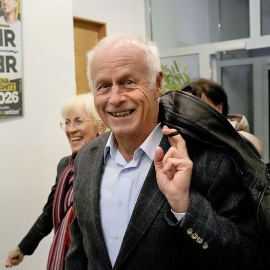 Ein älterer Mann mit weißem Haar lächelt und hält einen Mantel, steht neben einem Plakat. Eine Frau mit Brille und einem Schal steht hinter ihm.