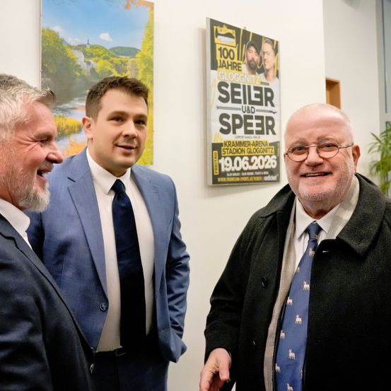Drei Männer in Anzügen stehen vor einer Wand. Zwei von ihnen lächeln, und der dritte schaut zur Kamera. Ein Plakat hinter ihnen bewirbt eine Gloggnitzer-Veranstaltung.