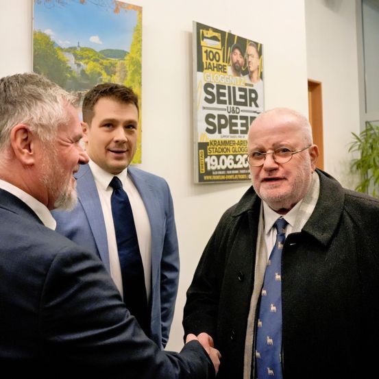Drei Männer in Anzügen stehen vor einem Plakat, wobei der Mann auf der linken Seite dem Mann in der Mitte die Hand schüttelt.
