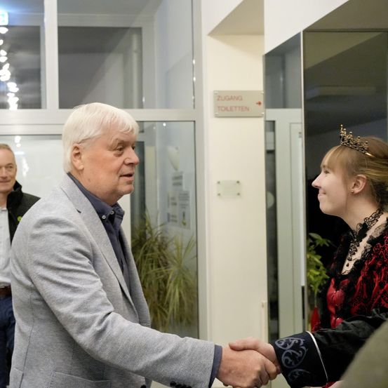 Ein älterer Mann mit weißen Haaren gibt einer Frau in einem rot-schwarzen Kleid die Hand. Hinter ihnen steht ein Mann in einem schwarzen Jackett. Der Ort ist innen mit Glastüren und einem Schild, das 'Zugang Toiletten' besagt.