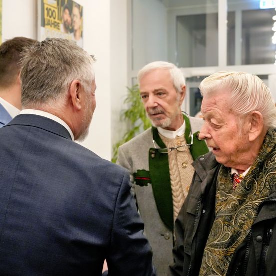 Drei Männer in formeller Kleidung unterhalten sich drinnen. Ein Mann im Anzug schaut einen Mann in einem grünen Pullover an. Hinter ihnen steht ein Mann mit einem gemusterten Schal.