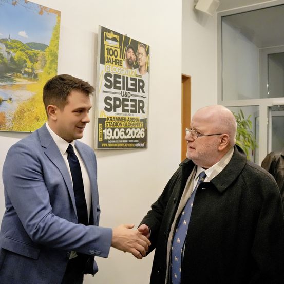 Zwei Männer in Anzughose und Krawatte schütteln sich die Hände vor einem Plakat für ein Konzert von Seiler und Speer. Das Plakat zeigt einen Mann mit Bart und das Datum des Konzerts.
