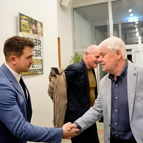 Zwei Männer schütteln sich die Hände in einem Flur, während ein dritter Mann hinter ihnen steht. Der Raum hat eine Glastür, eine Pflanze und ein Schild, das ein 100-jähriges Jubiläumsereignis ankündigt.