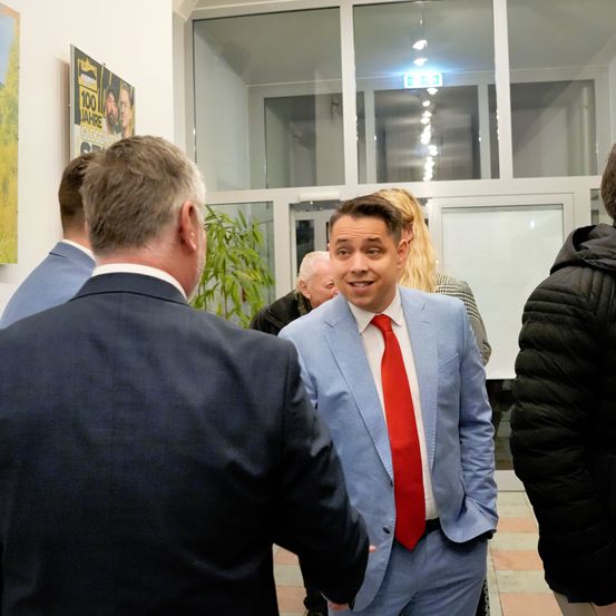 Mehrere Männer in Anzügen, darunter einer mit roter Krawatte, unterhalten sich in einem Raum mit Glaswänden und einem Poster mit der Aufschrift '100 Jahre' an der Wand.