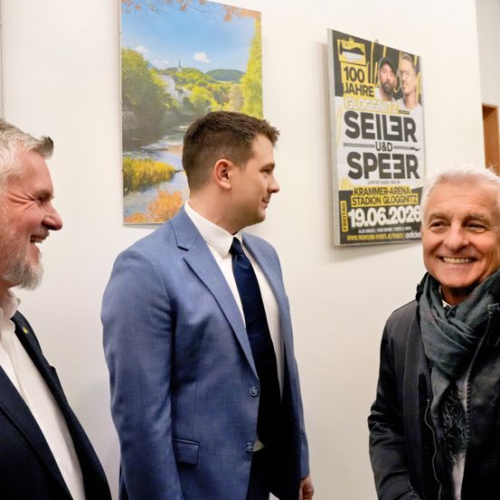 Drei Männer in Anzügen, einer mit Schal, lächelnd, vor einem Plakat mit 'Seiler und Speer' und einem Datum.