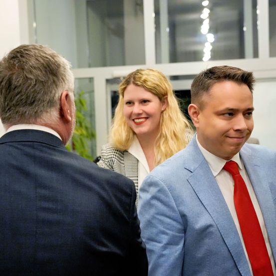 Drei Personen, zwei Männer und eine Frau, formell gekleidet, unterhalten sich in einem Raum mit Glasfenstern und einer Pflanze.