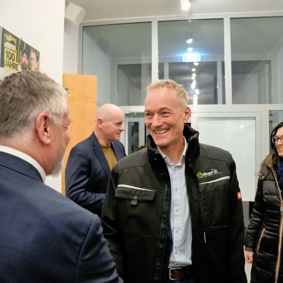 Drei Männer und eine Frau in einem Flur, sie lächeln. Ein Mann trägt eine schwarze Jacke mit Logo, ein anderer einen Anzug. Dahinter reflektiert eine Glaswand Lichter.