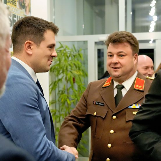 Zwei Männer, einer in einem Anzug und der andere in einer Militäruniform, schütteln sich die Hände in einer formellen Umgebung. Hinter ihnen beobachten zwei weitere Personen, eine ebenfalls in einem Anzug. Die Szene spielt sich in einem Raum mit einer Glaswand und Zimmerpflanzen ab.