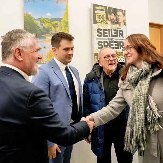 Vier Personen schütteln sich die Hände in einem Raum mit Plakaten im Hintergrund. Die Frau rechts lächelt und trägt eine Brille und einen Schal. Der Mann links trägt einen Anzug und eine Krawatte.