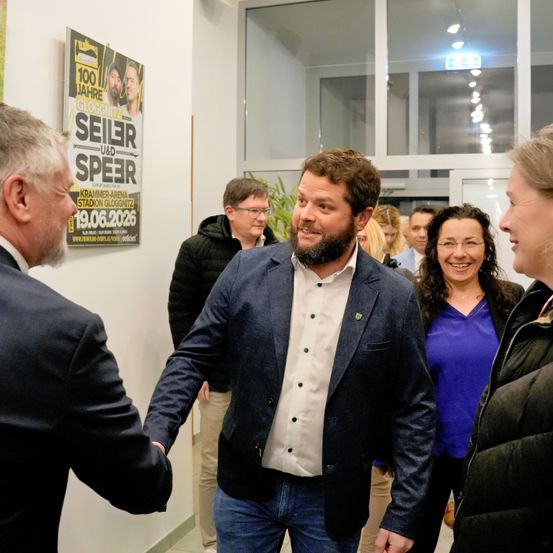 Eine Gruppe von Menschen befindet sich in einem Flur. Ein Mann im Anzug schüttelt einem anderen Mann in einem blauen Blazer und einem weißen Hemd die Hand. In der Nähe stehen eine Frau in einem blauen Shirt und eine andere in einer schwarzen Jacke und unterhalten sich. Ein Poster ist an der Wand angebracht.