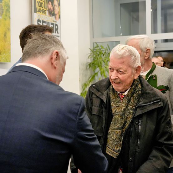 Mehrere Personen in formeller Kleidung sind drinnen versammelt, wobei ein älterer Mann lächelt und einem anderen Mann im Anzug die Hand schüttelt.