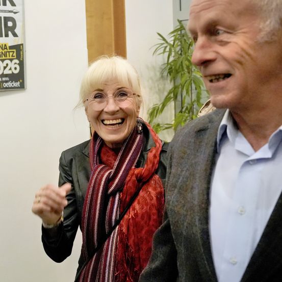 Eine ältere Frau mit blondem Haar, die eine Brille und einen roten Schal trägt, lächelt neben einem älteren Mann in Anzug und weißem Hemd. Hinter ihnen ist eine Wand mit einem Plakat.