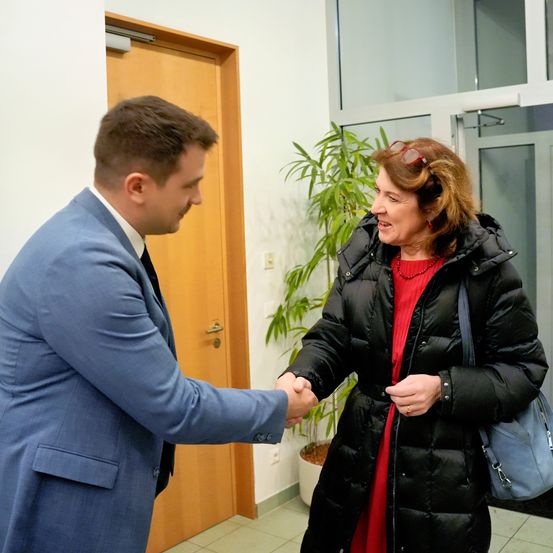 Ein Mann in einem blauen Anzug schüttelt die Hand einer Frau in einem schwarzen Mantel und einem roten Oberteil, die in einem Flur mit einer Holztür und einem Topf stehen.