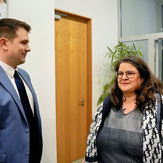 Ein Mann und eine Frau stehen nebeneinander in einem Büroflur, lächeln und schauen nach vorne.