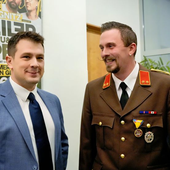 Zwei Männer stehen nebeneinander und lächeln. Ein Mann trägt eine Militäruniform mit Medaillen und einem Schnurrbart. Der andere Mann trägt einen Anzug.