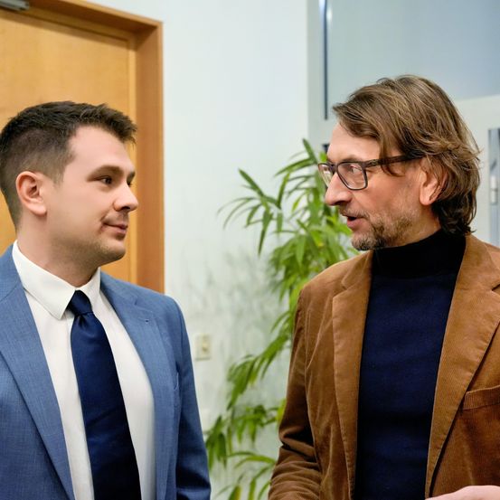 Zwei Männer stehen in einem Büro. Der Mann auf der linken Seite trägt einen blauen Anzug und eine Krawatte. Der Mann auf der rechten Seite trägt einen braunen Mantel, eine Brille und einen Rollkragenpullover.