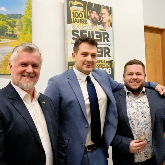 Drei Männer stehen lächelnd zusammen. Ein Mann trägt ein weißes Hemd, ein anderer ein Hemd mit Blumenmuster und der Mann in der Mitte einen blauen Anzug mit Krawatte. Hinter ihnen befindet sich ein Plakat für Gloggnitz.
