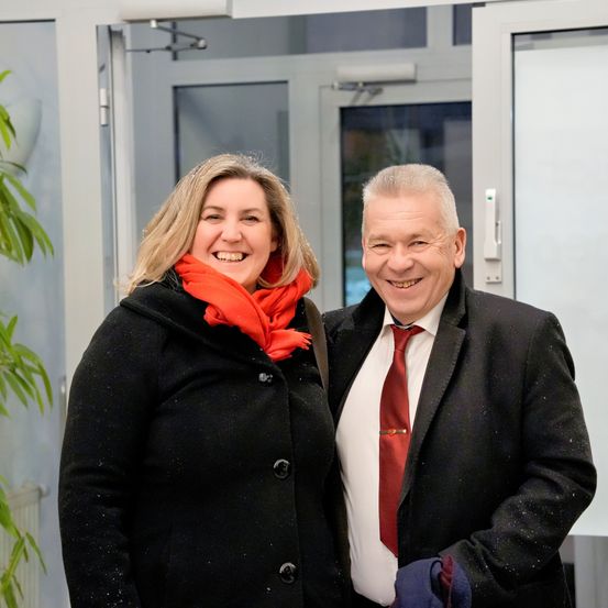 Ein Mann und eine Frau stehen nah beieinander draußen, beide tragen schwarze Mäntel und lächeln. Die Frau hat ein rotes Tuch. Hinter ihnen ist eine Glastür.