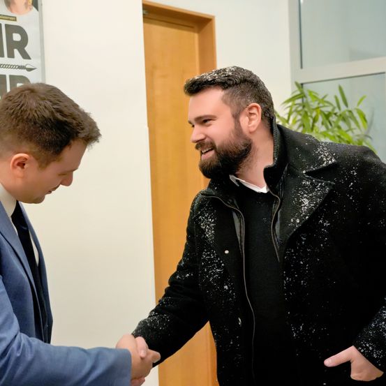 Zwei Männer schütteln sich die Hände in einem Innenbereich. Der Mann auf der linken Seite trägt einen blauen Anzug, während der Mann auf der rechten Seite einen schwarzen Mantel mit Schnee darauf hat. Hinter ihnen befindet sich eine Holztür und ein Poster mit weißem Text.