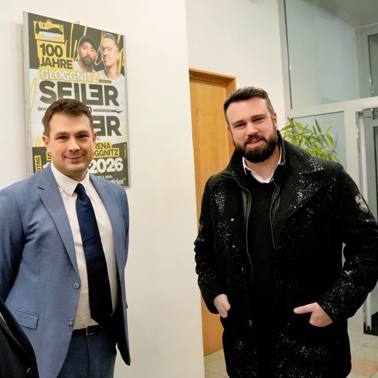 Zwei Männer stehen in einem Raum, einer trägt einen Anzug und der andere einen schwarzen Mantel mit weißen Sprenkeln. Hinter ihnen befindet sich ein Plakat für eine Gloggnitzer Veranstaltung.